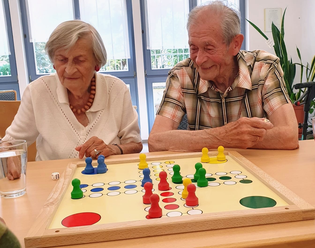 zwei ältere Menschen sitzen am Tisch. Vor ihnen liegt ein Brettspiel mit mehreren Figuren